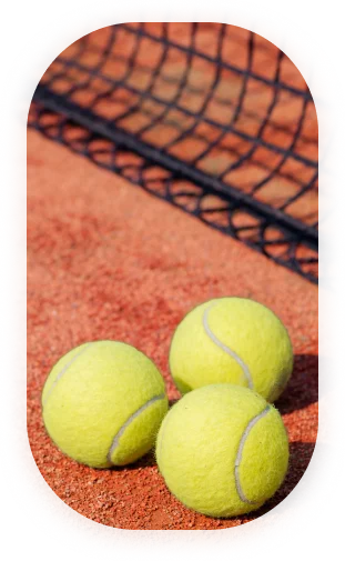 Trois balles de tennis jaunes reposent sur un court en terre battue, près d'un filet noir, projetant de légères ombres. La scène évoque une journée calme et ensoleillée sur un court de tennis