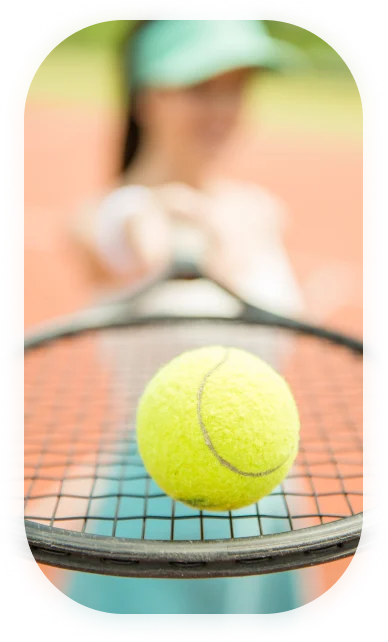 Gros plan sur une balle de tennis jaune posée en équilibre sur une raquette, tenue par une personne vêtue d'une tenue bleue et d'une visière sur un court de tennis ensoleillé, illustrant la concentration