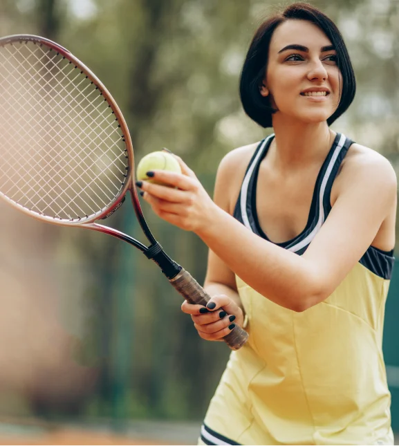 Une femme vêtue d'une robe de tennis jaune sourit alors qu'elle se prépare à servir. Elle tient une raquette et une balle de tennis sur un court en terre battue, avec des arbres en arrière-plan.